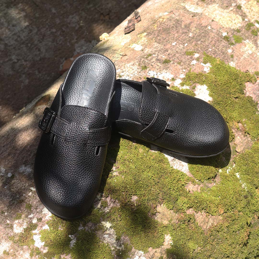 A pair of Thomas Chan's closed-toe buckle sandals in black colour, resting on a moss-covered log in an outdoor forest setting. The sandals feature a sleek design with a black buckle strap across the top and a black inner lining, with arch-support footbed.