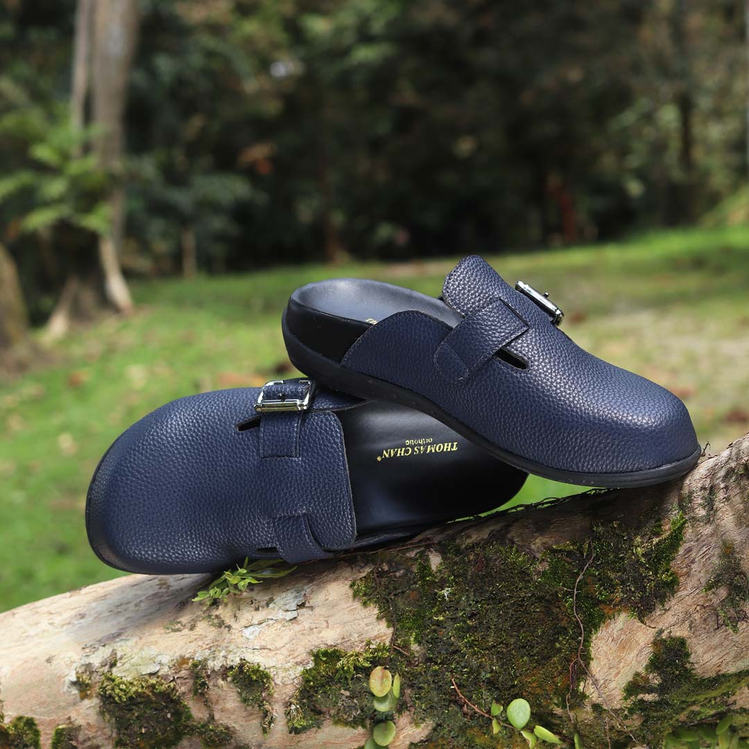 A pair of Thomas Chan's closed-toe buckle sandals in blue color, resting on a moss-covered log in an outdoor forest setting. The sandals feature a sleek design with a silver buckle strap across the top and a black inner lining, with arch-support footbed.