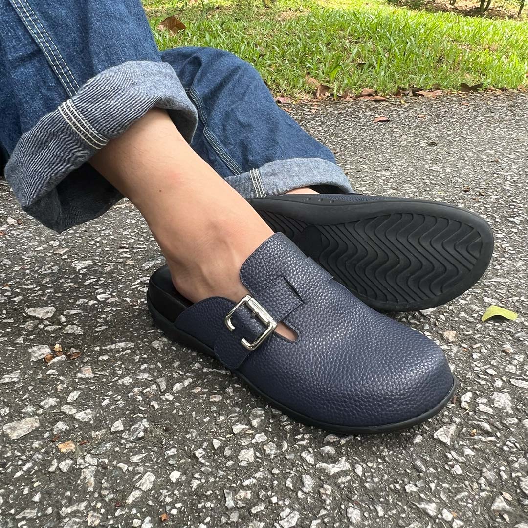 Person wearing rolled-up jeans and a pair of blue-coloured Thomas Chan's closed-toe buckle sandals while sitting on a paved surface. The sandals feature a sleek design with a silver buckle strap and with arch-support footbed.