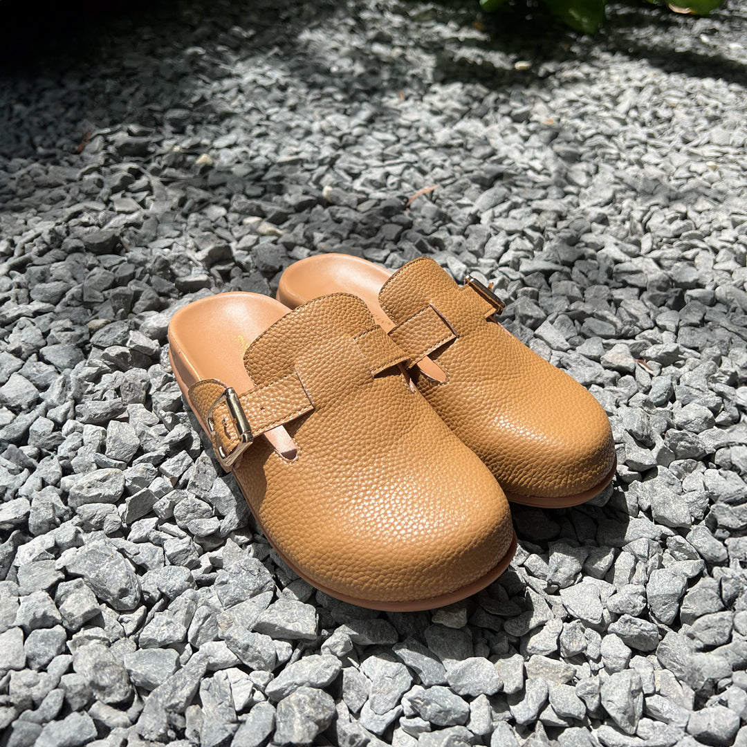 A pair of Thomas Chan's closed-toe buckle sandals in tan color, resting on a rocky surface. The sandals feature a sleek design with a gold buckle strap across the top and a foam inner lining, with an arch-support footbed.