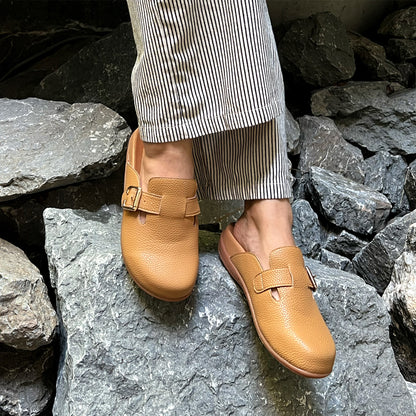 A pair of tan-colored closed-toe buckle sandals worn by a person standing on rocky terrain. The shoes feature a textured leather finish, a metal buckle detail, and are paired with striped pants.