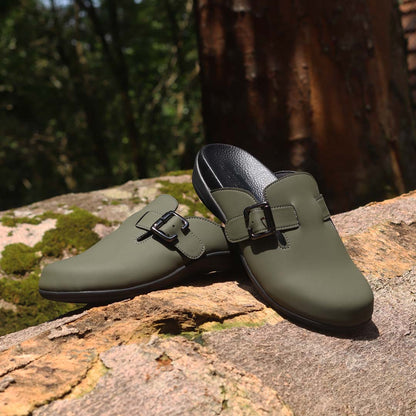 A pair of Thomas Chan's closed-toe buckle sandals in khaki color, resting on a moss-covered log in an outdoor forest setting. The sandals feature a sleek design with a black buckle strap across the top and a black inner lining, with arch-support footbed.