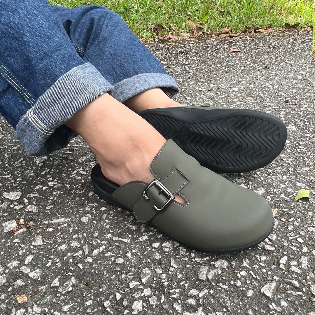 Person wearing rolled-up jeans and a pair of khaki-coloured Thomas Chan's closed-toe buckle sandals while sitting on a paved surface. The sandals feature a sleek design with a black buckle strap and with arch-support footbed.