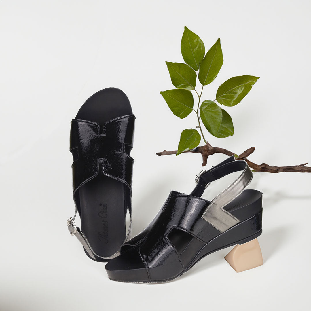 The image shows a pair of stylish black orthotic wedges. These shoes feature a unique H-strap with slingback design, providing both elegance and comfort. The wedges are from Thomas Chan Malaysia, as indicated by the branding inside the shoe. The setting is enhanced by the presence of a leaf in the light cream background, giving the image a casual and delicate feel.