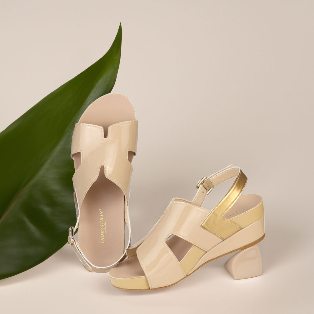 The image shows a pair of stylish cream orthotic wedges. These shoes feature a unique H-strap with slingback design, providing both elegance and comfort. The wedges are from Thomas Chan Malaysia, as indicated by the branding inside the shoe. The setting is enhanced by the presence of a leaf in the light cream background, giving the image a casual and delicate feel.