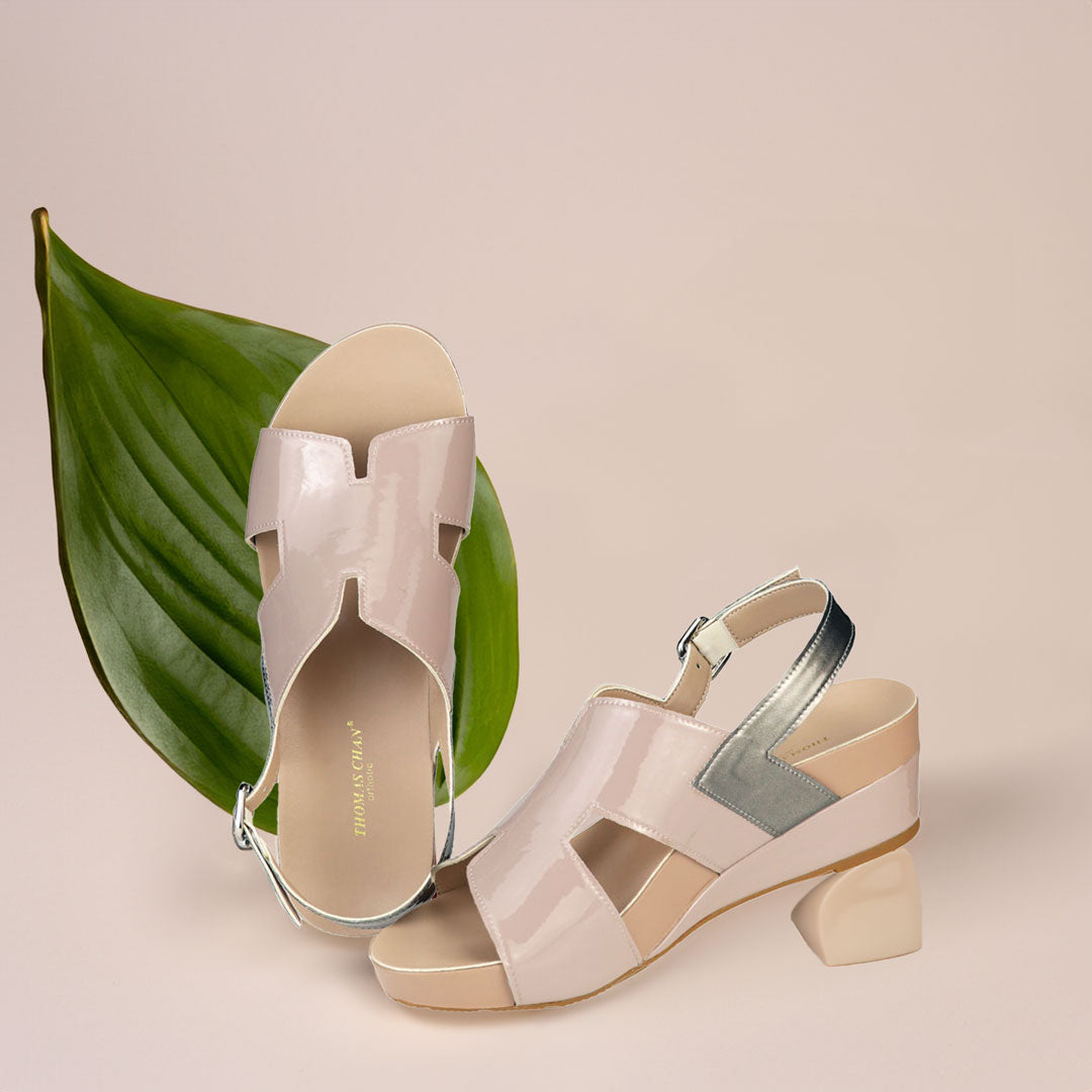 The image shows a pair of stylish light taupe orthotic wedges. These shoes feature a unique H-strap with slingback design, providing both elegance and comfort. The wedges are from Thomas Chan Malaysia, as indicated by the branding inside the shoe. The setting is enhanced by the presence of a leaf in the light cream background, giving the image a casual and delicate feel.