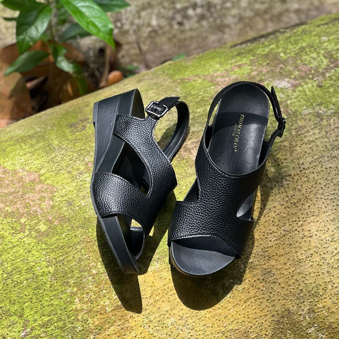 Thomas Chan Daily Wear Slingback Low Wedges in charcoal black displayed outdoors on a mossy surface. The sandals feature a textured pebbly lychee print, an elegant slingback design with a buckle closure, and a comfortable low wedge heel. The 'THOMAS CHAN®' brand is visible on the footbed.