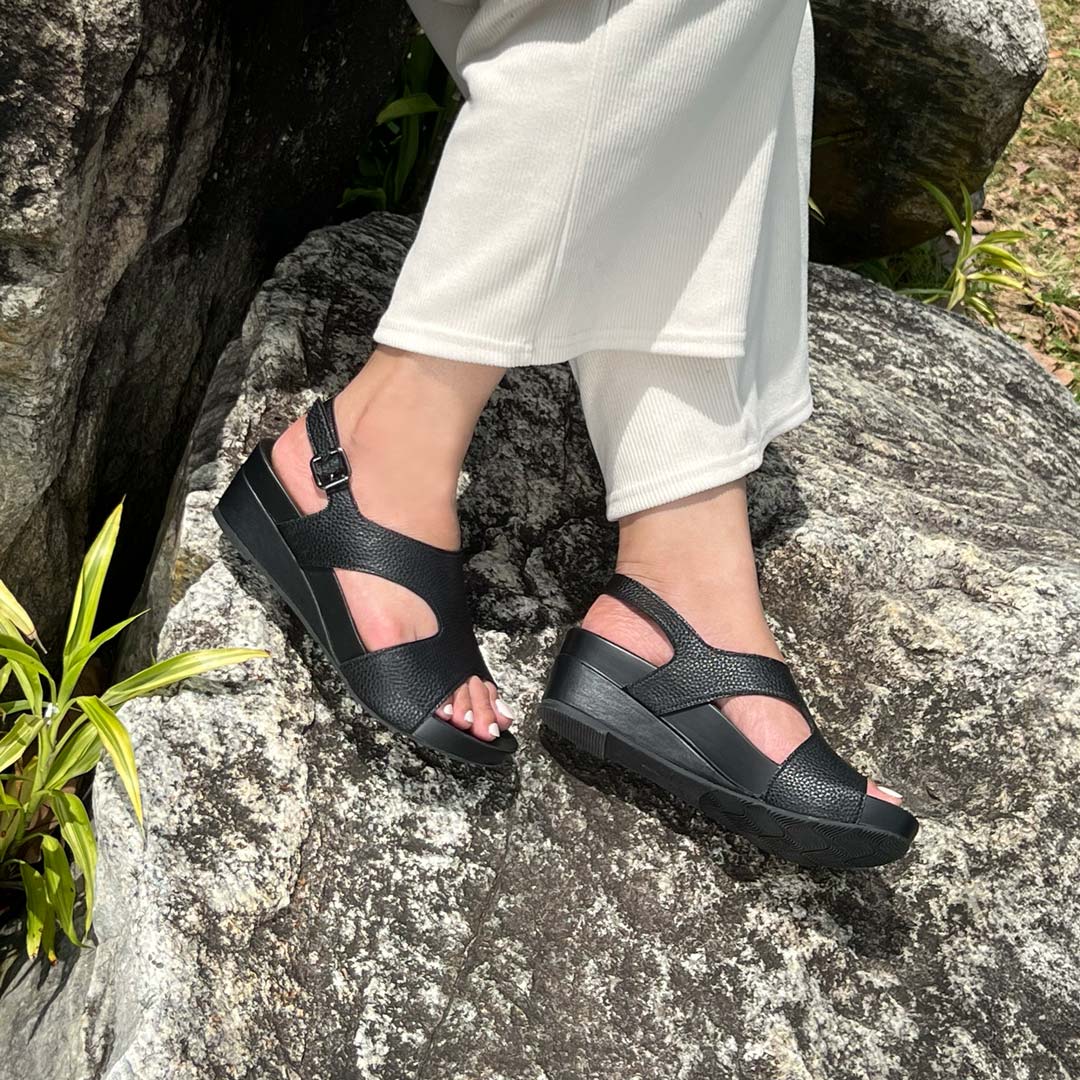 A model showcasting Thomas Chan Daily Wear Slingback Low Wedges in charcoal black outdoors on a rock surface. The sandals feature a textured pebbly lychee print, an elegant slingback design with a buckle closure, and a comfortable low wedge heel.