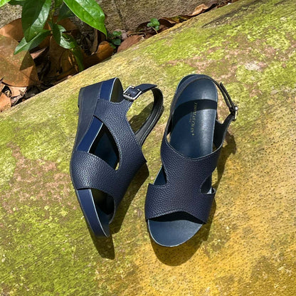 Thomas Chan Daily Wear Slingback Low Wedges in navy blue displayed outdoors on a mossy surface. The sandals feature a textured pebbly lychee print, an elegant slingback design with a buckle closure, and a comfortable low wedge heel. The 'THOMAS CHAN®' brand is visible on the footbed.