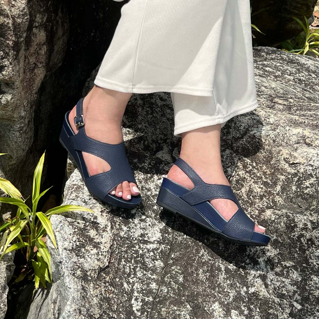 A model showcasting Thomas Chan Daily Wear Slingback Low Wedges in navy blue outdoors on a rock surface. The sandals feature a textured pebbly lychee print, an elegant slingback design with a buckle closure, and a comfortable low wedge heel.