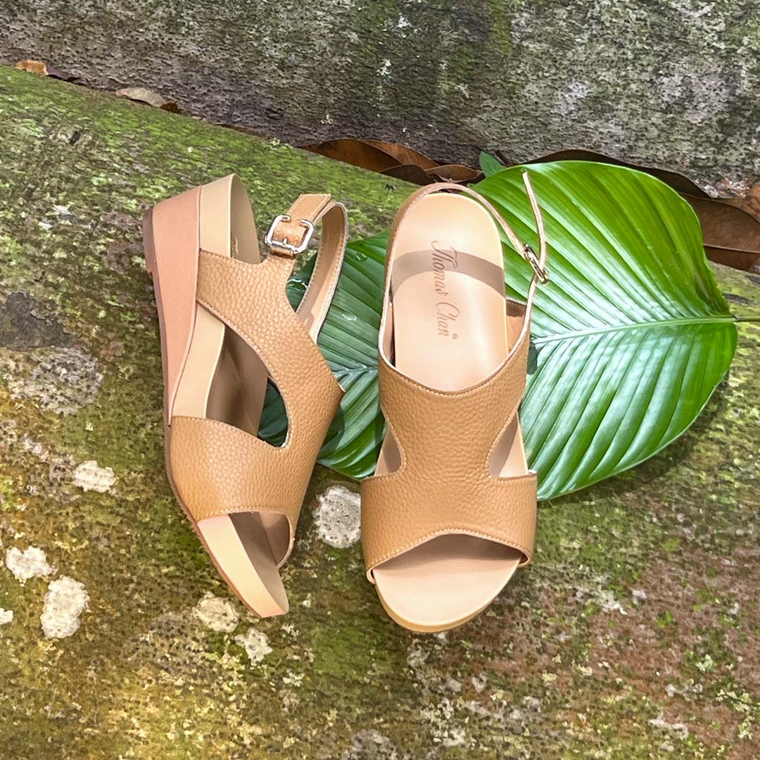 Thomas Chan Daily Wear Slingback Low Wedges in camel cream displayed outdoors under a leaf & mossy surface. The sandals feature a textured pebbly lychee print, an elegant slingback design with a buckle closure, and a comfortable low wedge heel. The 'THOMAS CHAN®' brand is visible on the footbed.