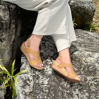 A model showcasting Thomas Chan Daily Wear Slingback Low Wedges in camel cream outdoors on a rock surface. The sandals feature a textured pebbly lychee print, an elegant slingback design with a buckle closure, and a comfortable low wedge heel.