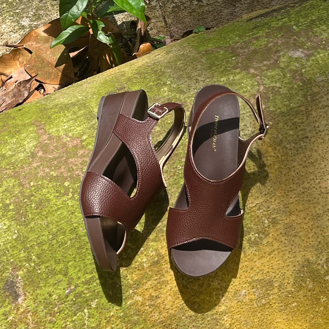 Thomas Chan Daily Wear Slingback Low Wedges in cocoa brown displayed outdoors on a mossy surface. The sandals feature a textured pebbly lychee print, an elegant slingback design with a buckle closure, and a comfortable low wedge heel. The 'THOMAS CHAN®' brand is visible on the footbed.