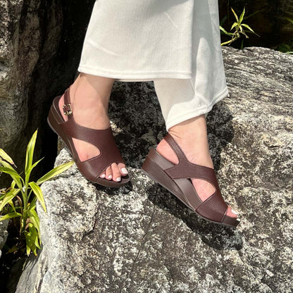 A model showcasting Thomas Chan Daily Wear Slingback Low Wedges in cocoa brown outdoors on a rock surface. The sandals feature a textured pebbly lychee print, an elegant slingback design with a buckle closure, and a comfortable low wedge heel.