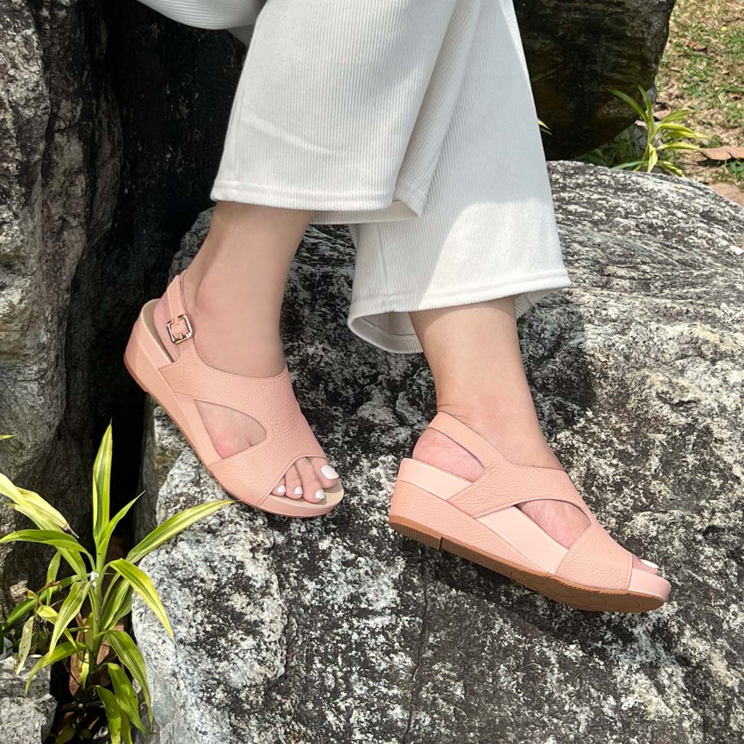 A model showcasting Thomas Chan Daily Wear Slingback Low Wedges in rosy pink outdoors on a rock surface. The sandals feature a textured pebbly lychee print, an elegant slingback design with a buckle closure, and a comfortable low wedge heel.