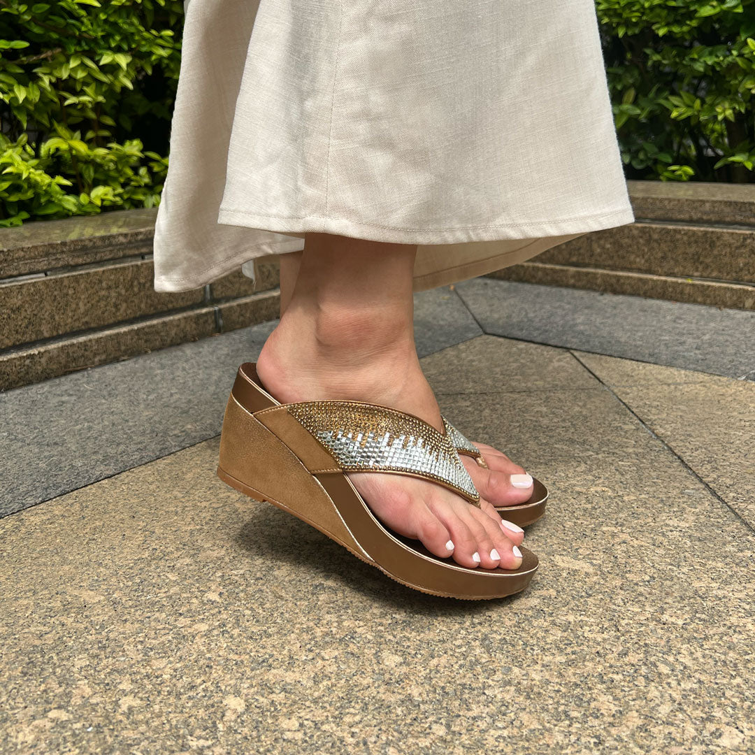 Model wearing Suede Rhinestone Wedge Flip Flops in bronze. Featuring a 3-inch wedge heel, these sandals provide exceptional comfort with the integrated Thomas Chan® Orthotic Arch-Legance footbed. Standing on rock tiles in the garden.