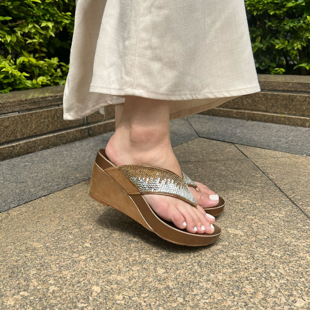 Model wearing Suede Rhinestone Wedge Flip Flops in bronze. Featuring a 3-inch wedge heel, these sandals provide exceptional comfort with the integrated Thomas Chan® Orthotic Arch-Legance footbed. Standing on rock tiles in the garden.