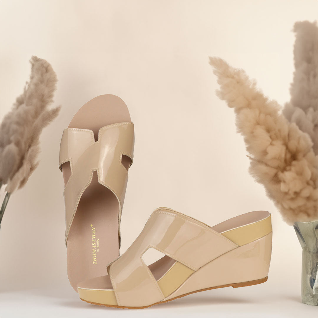 The image shows a pair of stylish cream orthotic wedges. These shoes feature a unique H-strap design, providing both elegance and comfort. The wedges are from Thomas Chan Malaysia, as indicated by the branding inside the shoe. The setting is enhanced by the presence of dried flowers in the background, giving the image a sophisticated and delicate feel.