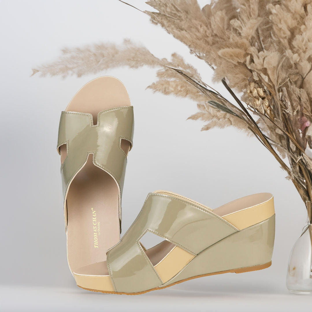 The image shows a pair of stylish ivory green orthotic wedges. These shoes feature a unique H-strap design, providing both elegance and comfort. The wedges are from Thomas Chan Malaysia, as indicated by the branding inside the shoe. The setting is enhanced by the presence of dried flowers in the background, giving the image a sophisticated and delicate feel.