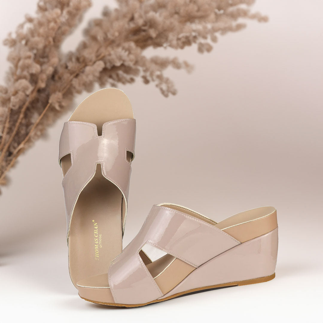 The image shows a pair of stylish light taupe orthotic wedges. These shoes feature a unique H-strap design, providing both elegance and comfort. The wedges are from Thomas Chan Malaysia, as indicated by the branding inside the shoe. The setting is enhanced by the presence of dried flowers in the background, giving the image a sophisticated and delicate feel.