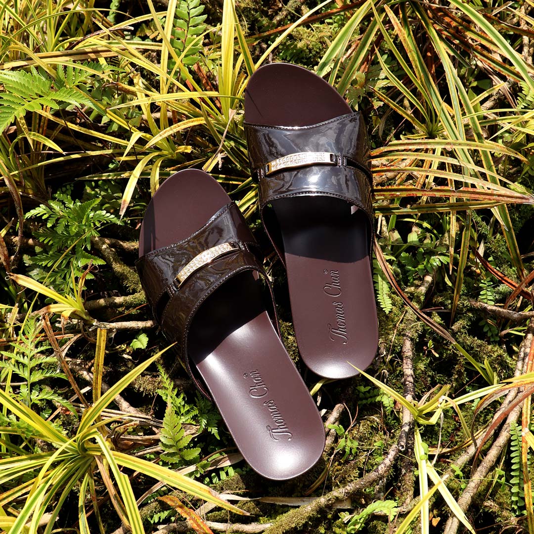 Thomas Chan Mini Charm Low Wedge Sandals in brown, featuring a delicate charm detail and a sleek coverage design. With a 2-inch wedge height and an arch-support footbed, these sandals provide a perfect blend of elegance and comfort. Placed against a backdrop of green and yellow plants, they evoke a classic yet laid-back vibe.