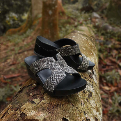 Thomas Chan's H-strap Glamour Stone Low Wedges in black colour, resting on a log in an outdoor forest setting. The sandals feature glamour rhinestone detailing ,and an arch-support footbed with 2-inch height wedges.