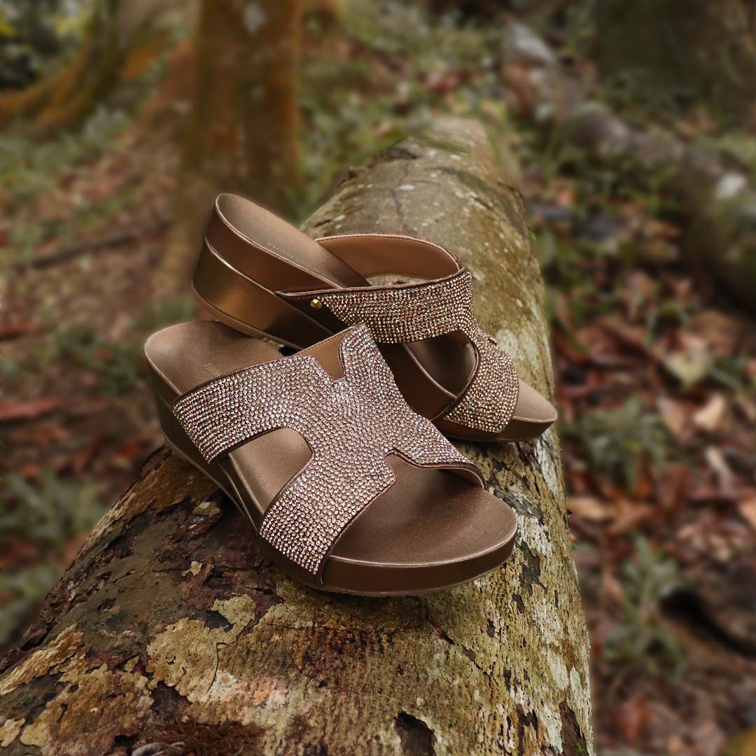 Thomas Chan's H-strap Glamour Stone Low Wedges in bronze colour, resting on a log in an outdoor forest setting. The sandals feature glamour rhinestone detailing ,and an arch-support footbed with 2-inch height wedges.