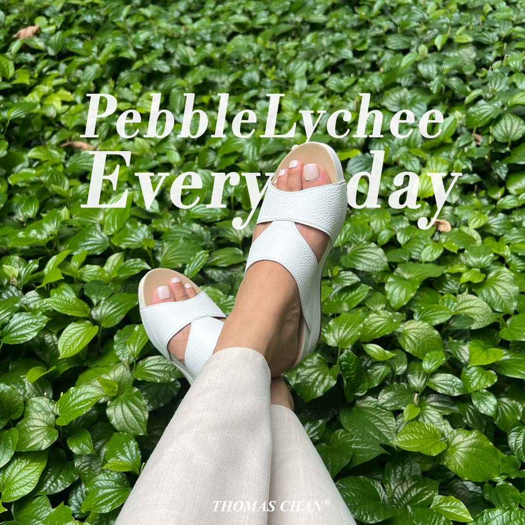 Model wearing Pebble Lychee Everyday Low Wedge Slides in pastel white. Featuring a 2-inch wedge heel, these sandals provide exceptional comfort with the integrated Thomas Chan® Orthotic Arch-Legance footbed. Green plants in the background.