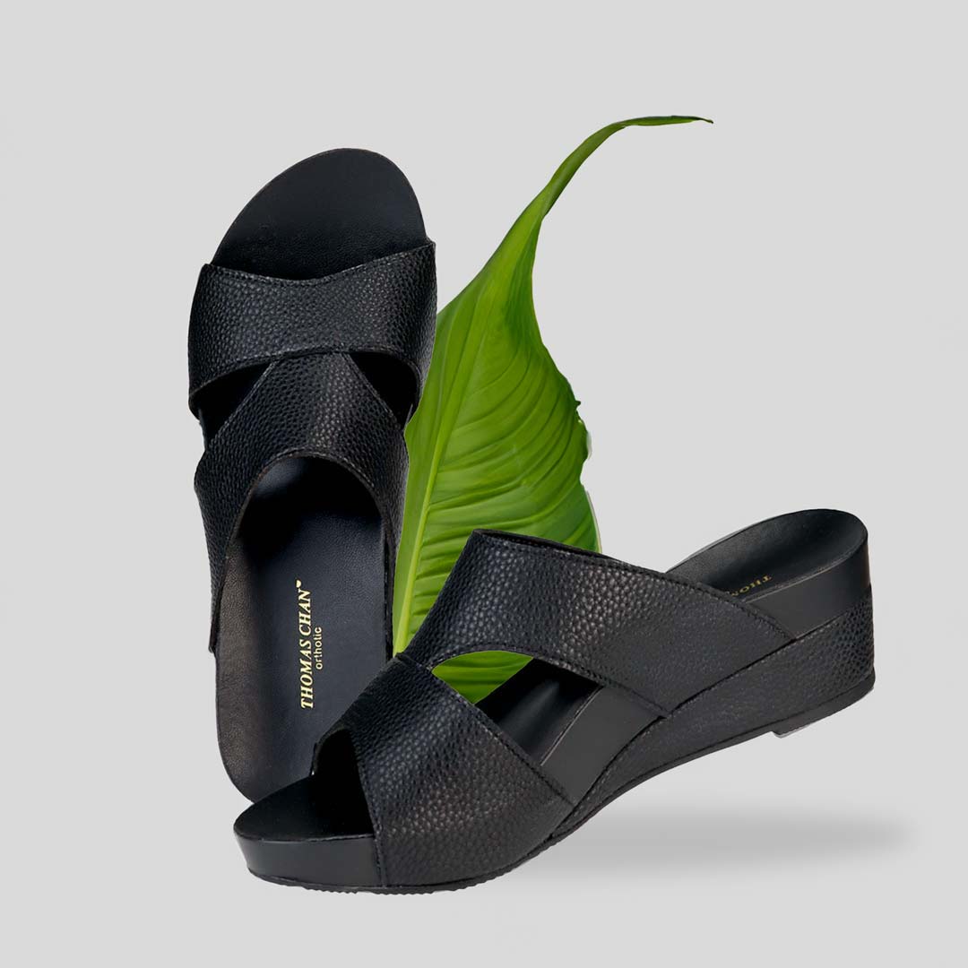 The image shows a pair of stylish Pebble Lychee Everyday Low Wedge Slides in black. These shoes feature a simple yet casual design, providing both elegance and comfort. The wedges are from Thomas Chan Malaysia, as indicated by the branding inside the shoe. The setting is enhanced by the presence of a leaf in the light cream background, giving the image a minimal and whimsical feel.