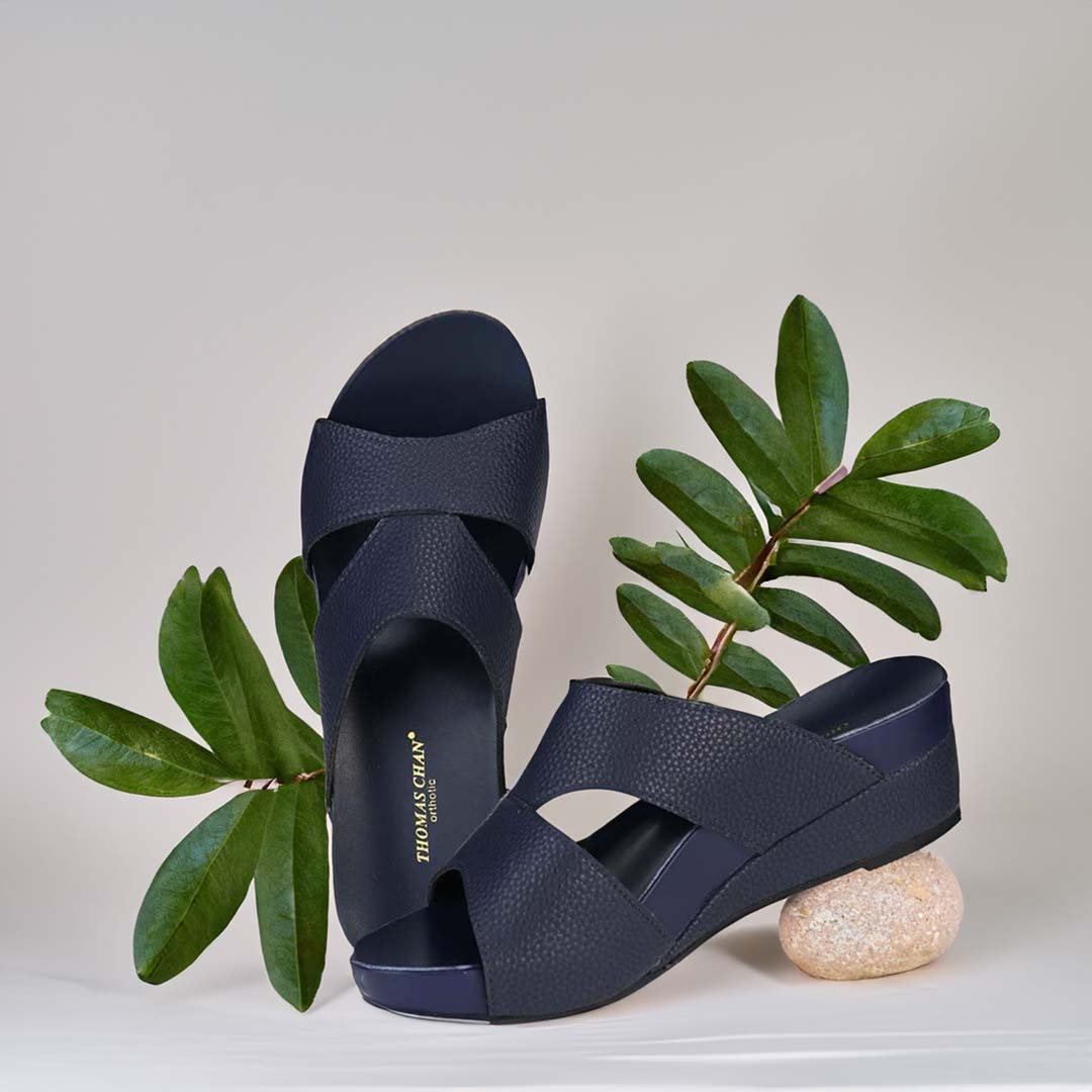 The image shows a pair of stylish Pebble Lychee Everyday Low Wedge Slides in dark blue. These shoes feature a simple yet casual design, providing both elegance and comfort. The wedges are from Thomas Chan Malaysia, as indicated by the branding inside the shoe. The setting is enhanced by the presence of leaves in the light beige background, giving the image a minimal and whimsical feel.