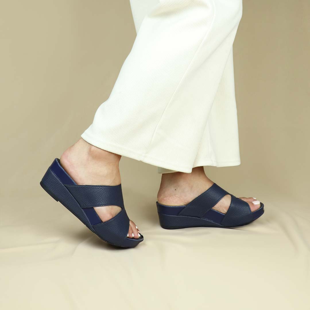 Model wearing Pebble Lychee Everyday Low Wedge Slides in dark blue. Featuring a 2-inch wedge heel, these sandals provide exceptional comfort with the integrated Thomas Chan® Orthotic Arch-Legance footbed.