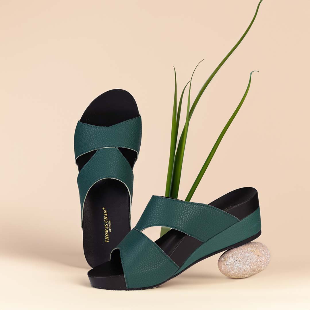 The image shows a pair of stylish Pebble Lychee Everyday Low Wedge Slides in dark green. These shoes feature a simple yet casual design, providing both elegance and comfort. The wedges are from Thomas Chan Malaysia, as indicated by the branding inside the shoe. The setting is enhanced by the presence of a leaf in the light cream background, giving the image a minimal and whimsical feel.