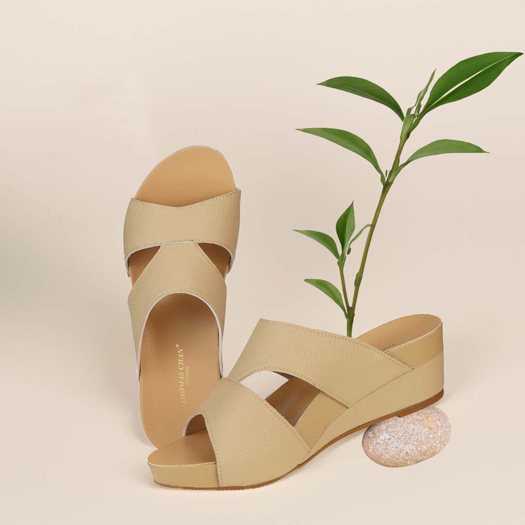 The image shows a pair of stylish Pebble Lychee Everyday Low Wedge Slides in mustard cream. These shoes feature a simple yet casual design, providing both elegance and comfort. The wedges are from Thomas Chan Malaysia, as indicated by the branding inside the shoe. The setting is enhanced by the presence of a leaf in the light cream background, giving the image a minimal and whimsical feel.