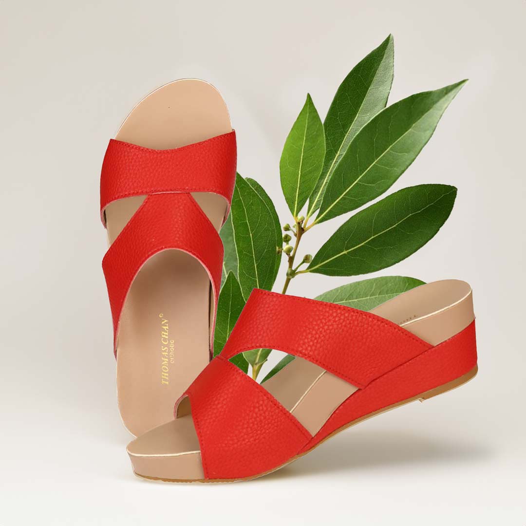 The image shows a pair of stylish Pebble Lychee Everyday Low Wedge Slides in red. These shoes feature a simple yet casual design, providing both elegance and comfort. The wedges are from Thomas Chan Malaysia, as indicated by the branding inside the shoe. The setting is enhanced by the presence of a leaf in the light cream background, giving the image a minimal and whimsical feel.