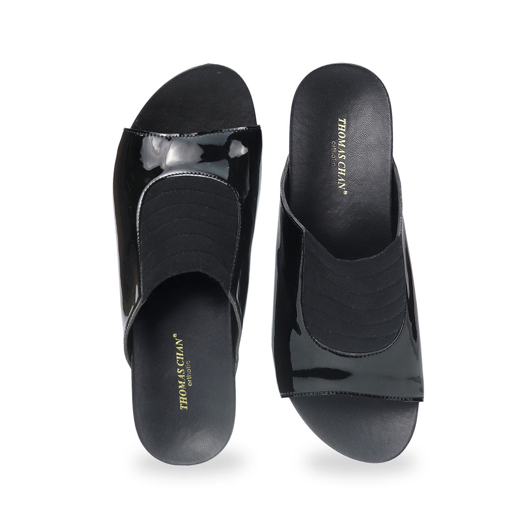 Front view of Thomas Chan's Matte Cushioned Low Wedge Sandals in black, highlighting the matte leather front padding, pattern leather accents, and signature arch-support footbed for all-day comfort and style. Set outdoors on a cement floor with lush grass in the background.