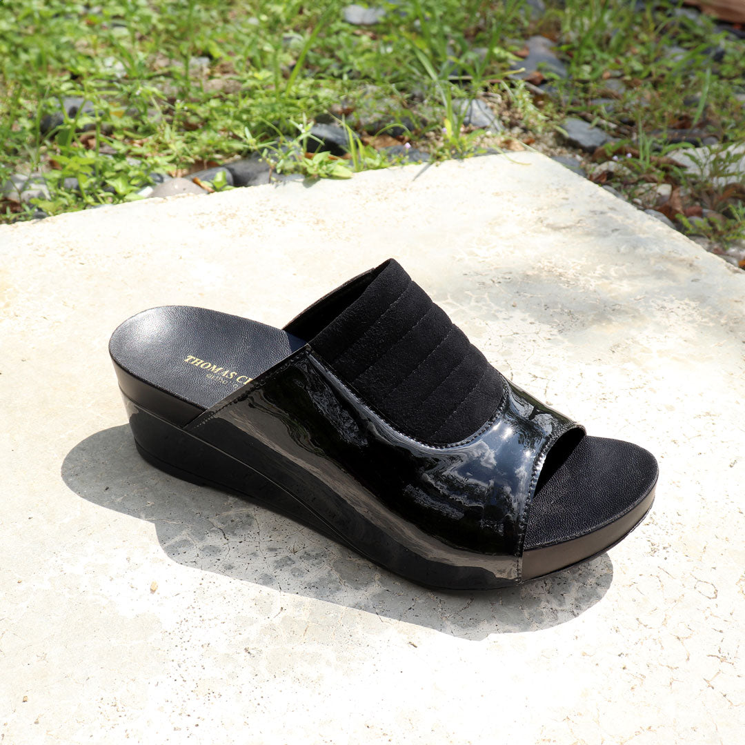 Diagonal view of Thomas Chan's Matte Cushioned Low Wedge Sandals in black, highlighting the matte leather front padding, pattern leather accents, and signature arch-support footbed for all-day comfort and style. Set outdoors on a cement floor with lush grass in the background.