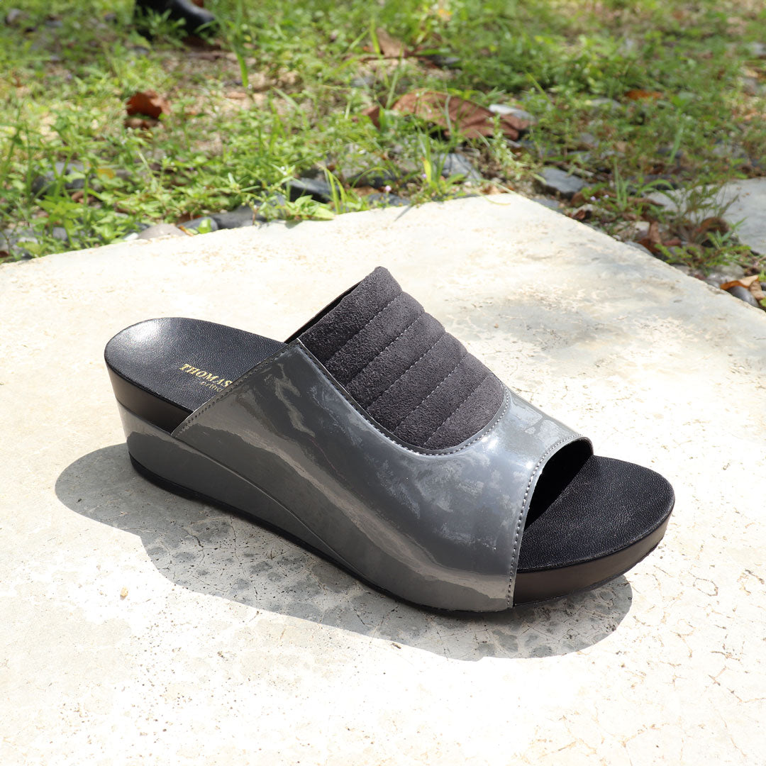 Diagonal view of Thomas Chan's Matte Cushioned Low Wedge Sandals in grey, highlighting the matte leather front padding, pattern leather accents, and signature arch-support footbed for all-day comfort and style. Set outdoors on a cement floor with lush grass in the background.