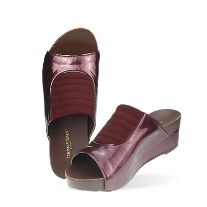 Full view of Thomas Chan's Matte Cushioned Low Wedge Sandals in maroon, highlighting the matte leather front padding, pattern leather accents, and signature arch-support footbed for all-day comfort and style. Set outdoors on a cement floor with lush grass in the background.