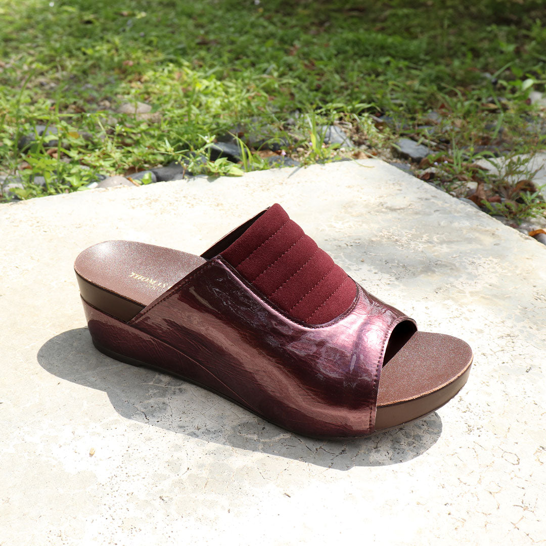 Diagonal view of Thomas Chan's Matte Cushioned Low Wedge Sandals in maroon, highlighting the matte leather front padding, pattern leather accents, and signature arch-support footbed for all-day comfort and style. Set outdoors on a cement floor with lush grass in the background.