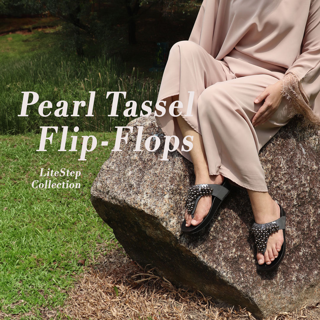 A person is seated on a rock outdoors wearing Thomas Chan's Pearl Tassel Flip-Flops from the LiteStep Collection. The sandals feature a glossy dark strap adorned with pearl tassel embellishments, paired with a comfortable flatform sole. The outfit complements the footwear with a flowy beige dress with lace details on the sleeves. The natural outdoor setting enhances the relaxed yet elegant vibe of the sandals. The "THOMAS CHAN®" branding and product description are displayed in the image.