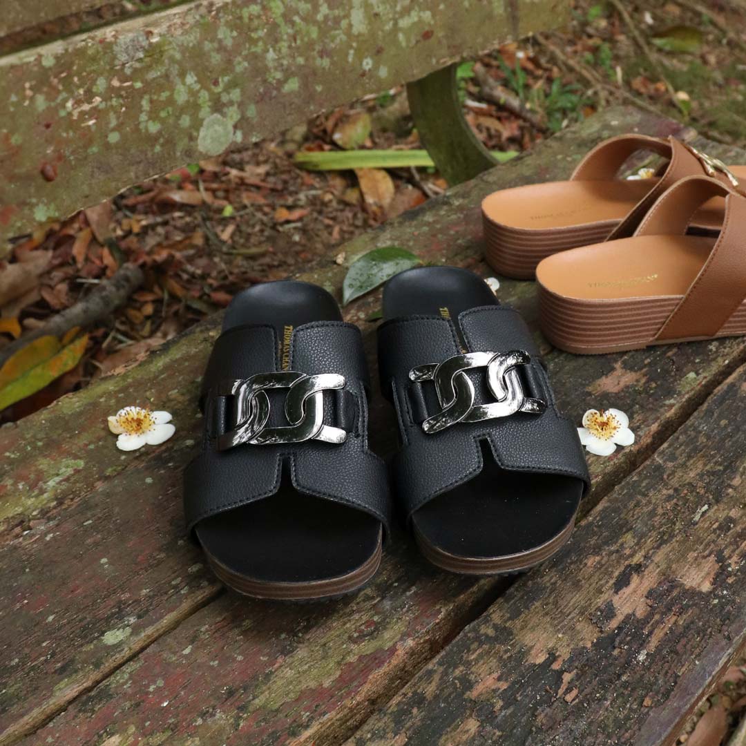 Thomas Chan's Metallic Buckle H-Flatform Sandals in black, featuring a bold metallic buckle across the H-strap and LiteStep flatform footbed, displayed outdoors on a weathered wooden bench with floral accents.