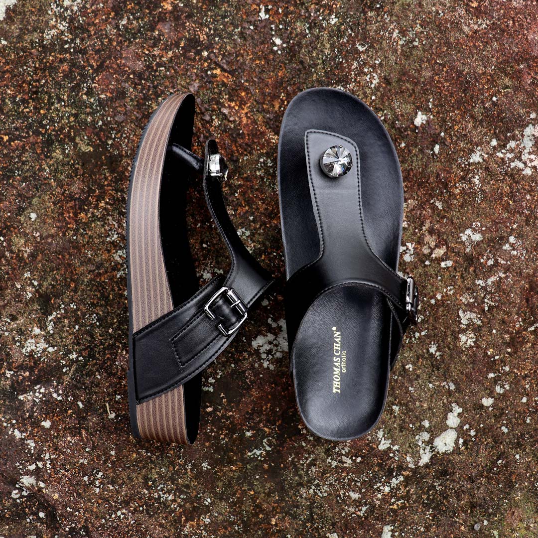 Full view of Thomas Chan’s Dazzle Crystal Adjustable Flip-Flops in black, placed on a red stone outdoors. The image showcases the sparkling crystal stone, adjustable buckle strap, and LiteStep flatform footbed, emphasizing their casual yet elegant style.