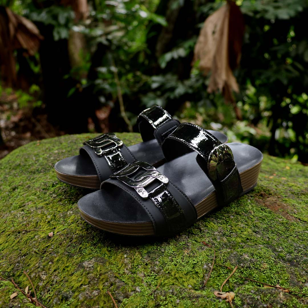 Full diagonal view of Thomas Chan’s Metallic Duo-Strap Adjustable Sandals in black, placed on a green moss-covered stone outdoors. The image highlights sleek metallic straps with a decorative buckle at the front, an adjustable instep buckle strap for a customizable fit, and a LiteStep flatform footbed, emphasizing their casual yet elegant style.