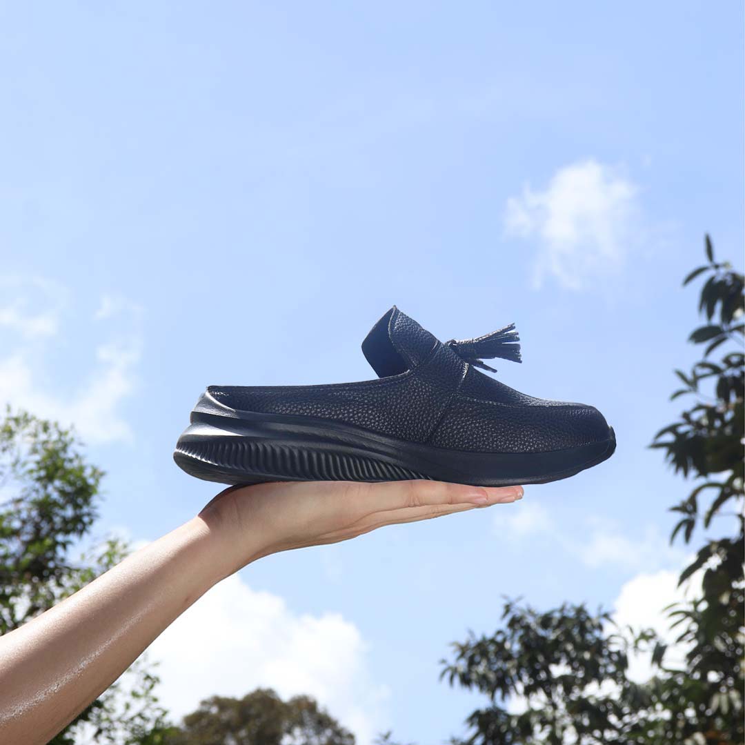  A hand holding Thomas Chan Feather-Light Tassel Slide-On Loafers in ivory, featuring a tassel bell design and a comfortable cushioned footbed, with a blue sky and leaves in the background