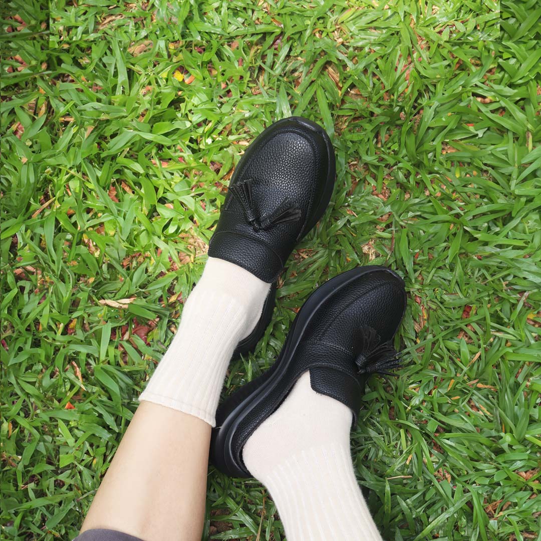 A model showcasing Thomas Chan Feather-Light Tassel Slide-On Loafers in black with stockings on, featuring a tassel bell design and a comfortable cushioned footbed, sitting on grass.