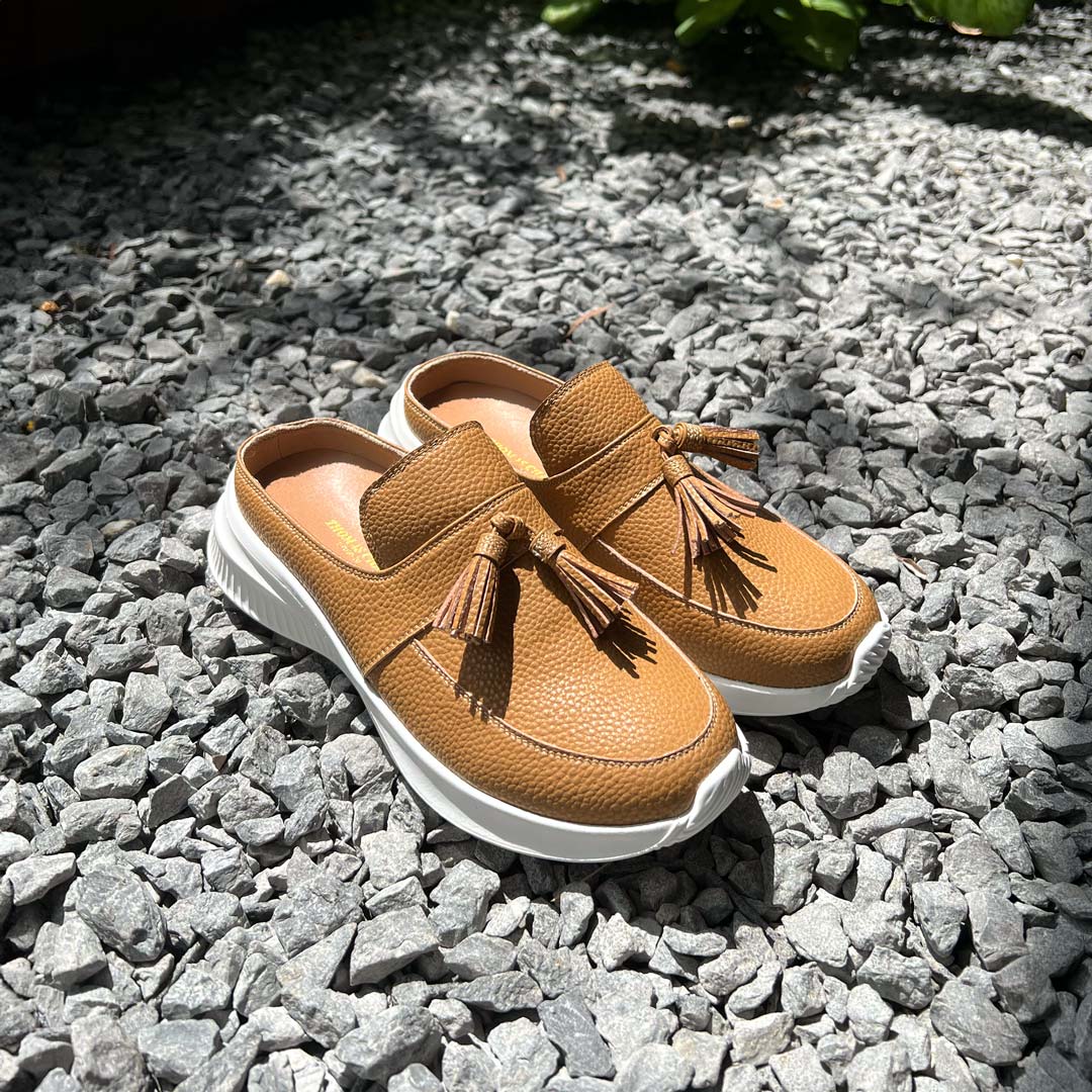 Thomas Chan Feather-Light Tassel Slide-On Loafers in camel, showcasing a stylish tassel bell design and cushioned footbed, displayed on a gravel stone surface under warm sunlight.