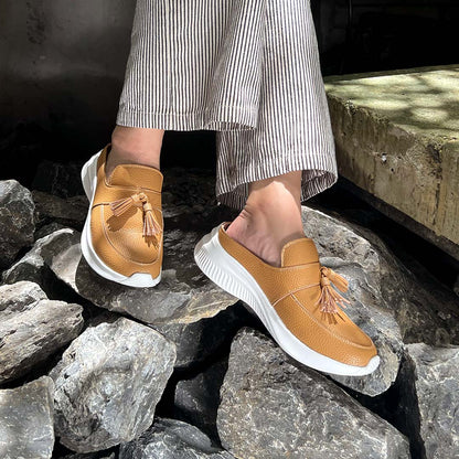 A model showcasing Thomas Chan Feather-Light Tassel Slide-On Loafers in camel, with a chic tassel bell design and cushioned footbed, seated on a lime-colored platform surrounded by large gravel stones.