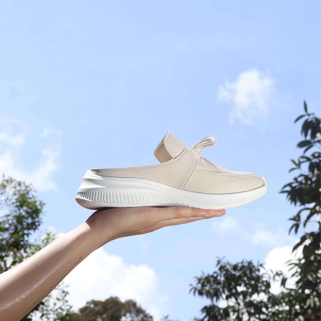 A hand holding Thomas Chan Feather-Light Tassel Slide-On Loafers in ivory, featuring a tassel bell design and a comfortable cushioned footbed, with a blue sky and leaves in the background