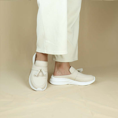 A model showcasing Thomas Chan Feather-Light Tassel Slide-On Loafers in ivory, featuring a tassel bell design and a comfortable cushioned footbed, standing against a cream backdrop.