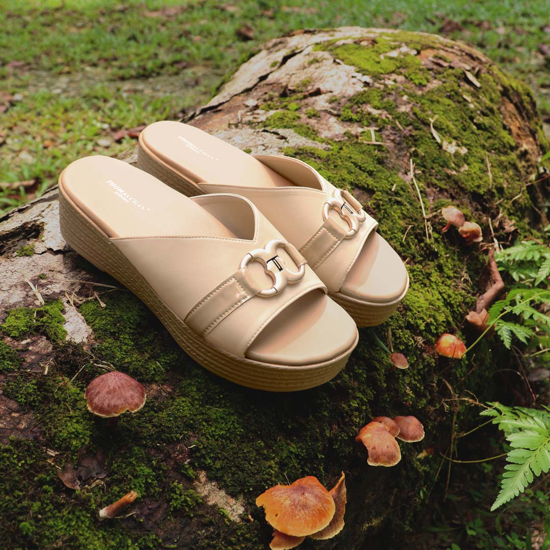 Diagonal view of the Thomas Chan Minimalist Buckle Flatform Sandals in cream, featuring a refined infinity buckle adorned with the signature 'T' logo. The cushioned footbed ensures all-day comfort, while the sandals are beautifully set against a serene forest backdrop, resting on a moss-covered surface dotted with mushrooms.